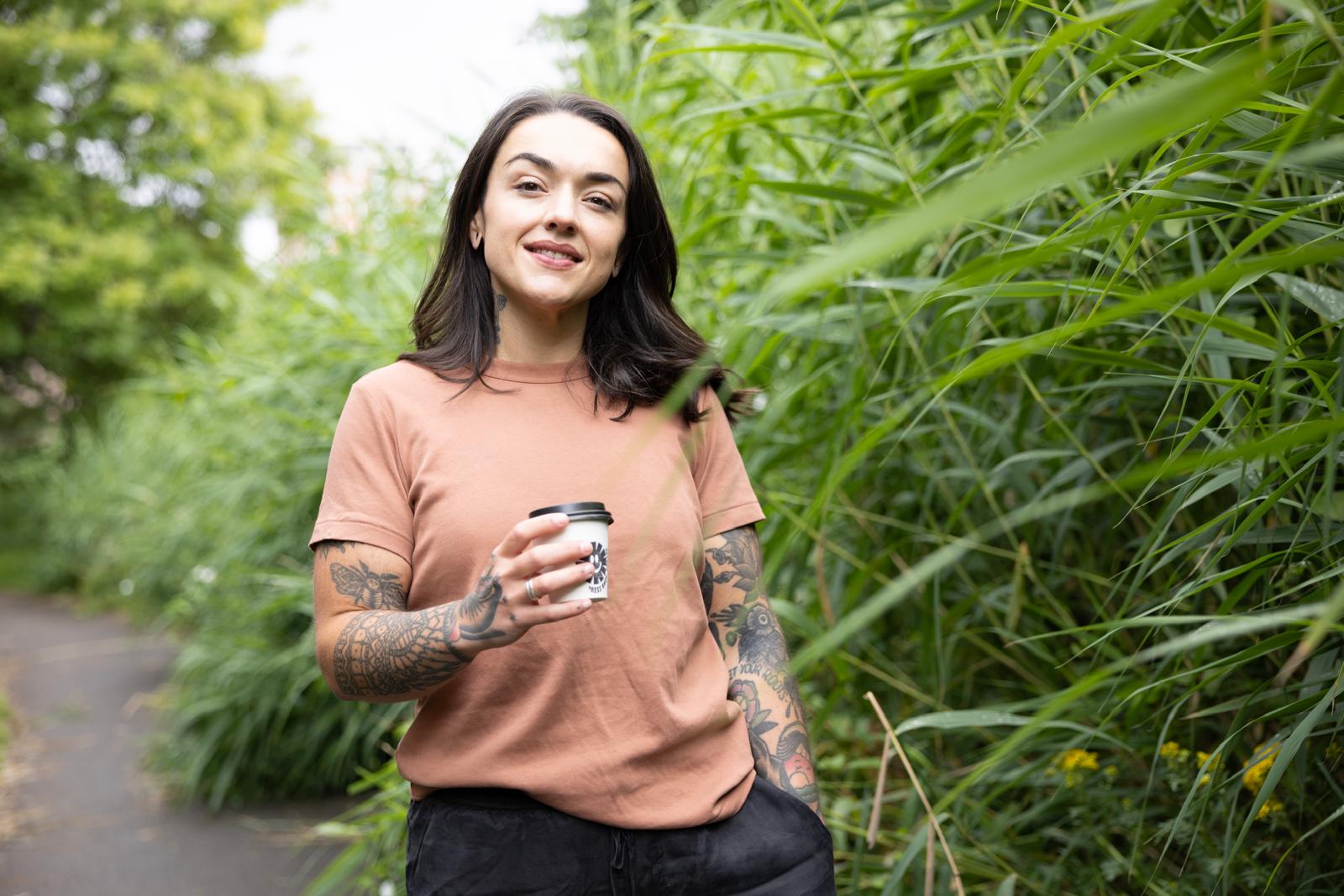 Alex Hindley standing on a path between tall green reeds, holding a coffee and looking at the camera